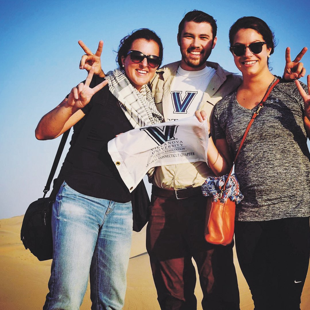 A group of three Villanova alumni holding a small white Villanova flag in the desert of Abu Dhabi, United Arab Emirates