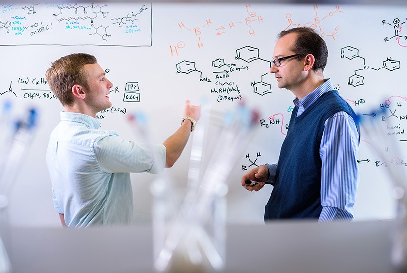 Undergraduate Programs in Chemistry image of student and professor discussing chemical equations at a whiteboard.