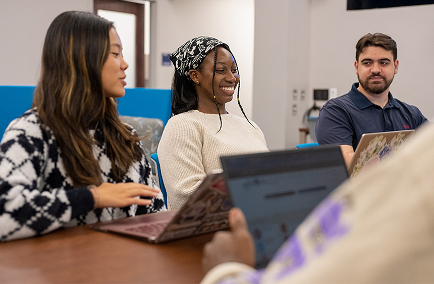 three students sit next to each other at a table with their laptops open in front of them