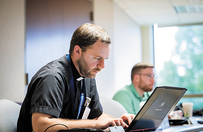 a priest wearing a clerical collar sits in a classroom looking at the screen of an open laptop